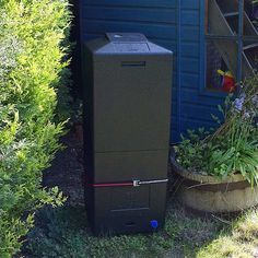 a tall black trash can sitting next to a blue building in the grass and bushes