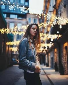 a young woman standing in an alleyway with lights strung all over the buildings behind her