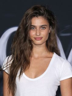 a woman with long brown hair and blue eyes wearing a white t - shirt is posing for the camera