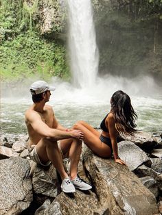 a man and woman sitting on rocks in front of a waterfall