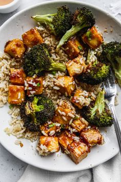 a white plate topped with tofu and broccoli covered in sauce next to rice