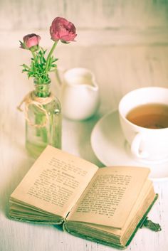 an open book sitting on top of a table next to a cup of tea and a vase