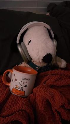 a teddy bear with headphones and a coffee mug on a bed next to a blanket