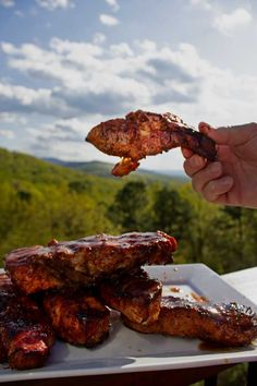 someone is holding up some meat on a plate with mountains in the backgroud