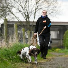 a woman walking her dog on a leash down a path in the grass near a fence