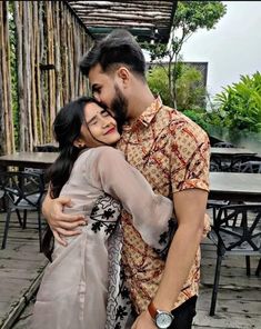 a man and woman embracing each other in front of a wooden structure with tables behind them