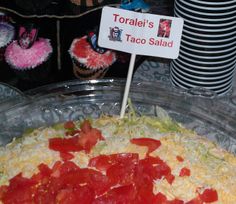 a taco salad with tomatoes and cheese in a glass dish on a table next to cupcakes