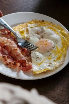 a person is holding a fork and knife over a plate with eggs and bacon on it