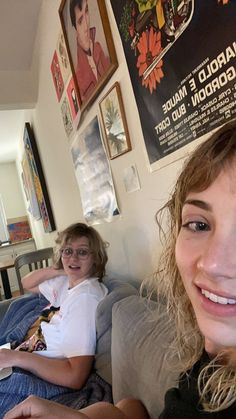 two young women sitting on a couch in a living room with posters above the couch