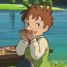 a young boy sitting at a table eating food in front of a body of water