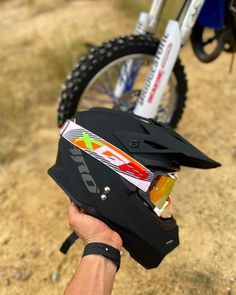 a person holding up a black helmet on top of a dirt road next to a bike