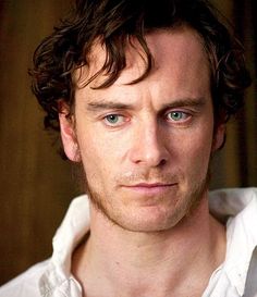 a man with curly hair and blue eyes looks at the camera while wearing a white shirt