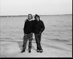 two young men standing on the beach next to the ocean with their arms around each other