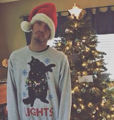 a man wearing a light up christmas sweater and santa hat standing in front of a decorated christmas tree