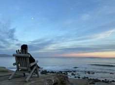 a person sitting on a wooden bench near the ocean at sunset or dawn with their back turned to the camera