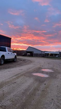 a white truck parked on top of a dirt road next to a barn at sunset