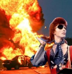 an image of a man in front of a fire with sunglasses on his head and a car behind him