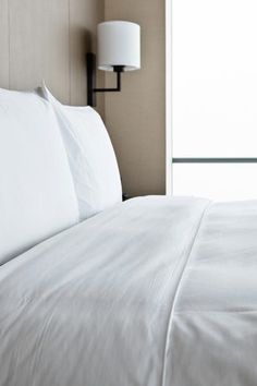 a bed with white sheets and pillows in a hotel room next to a lamp on the wall