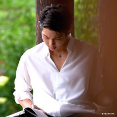 a man sitting down reading a book in front of a tree and looking at his phone