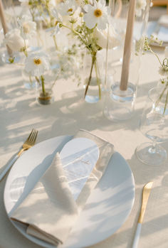 the table is set with white plates, silverware and flowers in glass vases