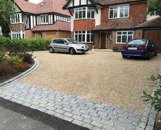 two cars are parked in front of a large brick driveway with trees and bushes on both sides