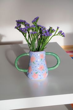 purple flowers are in a blue and pink vase on a table with a white wall behind it