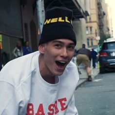 a young man in a white sweatshirt and black hat is laughing on the side walk