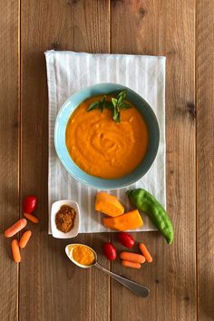 a bowl of carrot soup on a wooden table with other vegetables and spices around it