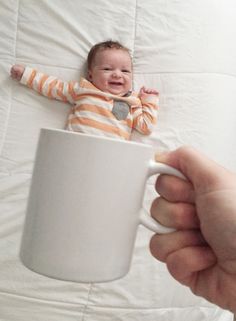 a person holding a coffee mug with a small baby in it's lap on a bed