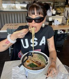 a woman eating noodles with chopsticks at a table in a restaurant, while wearing sunglasses