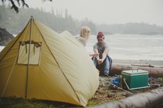 two people sitting on the ground next to a yellow tent and some logs near water