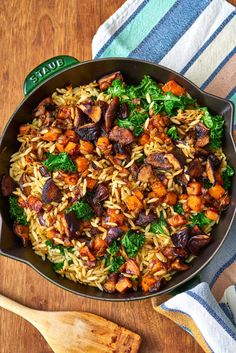 a skillet filled with rice and vegetables on top of a wooden table next to a spoon