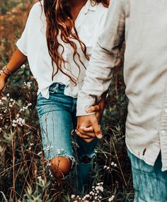 a man and woman holding hands walking through tall grass with wildflowers in the background