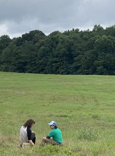 two people are sitting in the middle of a grassy field with trees in the background