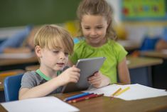 two young children sitting at desks playing on an electronic device in front of them