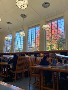 people are sitting at tables in a library with large windows and wooden chairs around them