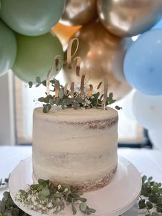 a white cake sitting on top of a table with balloons and greenery around it