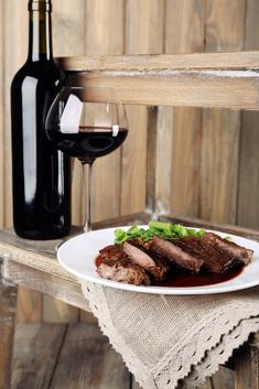 a white plate topped with meat next to a bottle of wine and a glass of red wine