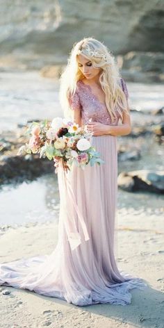 a beautiful blonde woman in a long dress holding a bouquet on the beach by the water