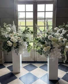 two white vases with flowers in them sitting on a checkered floor next to a window