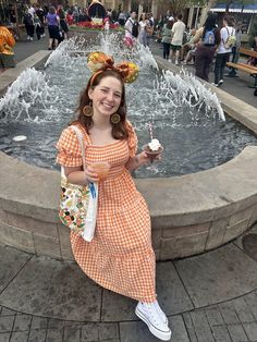 a woman in an orange and white checkered dress holding a cupcake near a fountain