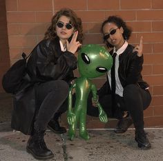 two people sitting next to each other near a brick wall with an alien doll in front of them