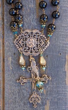 "BEADS: A stunning and bold combination of genuine high quality 10mm black onyx natural stones, with aqua and champagne crystal accents.    CENTERPIECE MEDALS: These are solid bronze hand cast designs featuring a 1.5\" tall design depicting Our Blessed Mother Mary on a beautiful filigree setting. The cross is a 2.25\" tall design detailing an elaborate cross of European origin.  ANGEL WING: Each design comes with the trademark Seraphym 1\" tall angel wing.      NECKLACE LENGTH AND SIZING: This design measures a total of 18\" not including the drop length of the medal. Each strand measures 9″ from the clasp located at the back of the neck to the top of the centerpiece medal. We can modify the length to your preference. Please indicate preferred length at check out. Additional cost to add le Elegant Bronze Jewelry With Patina, Luxury Engraved Bronze Jewelry, Luxury Collectible Bronze Jewelry, Black Spiritual Jewelry With Antique Finish, Black Brass Spiritual Jewelry, Spiritual Bronze Jewelry With Unique Variations, Ornate Bronze Jewelry With Decorative Features, Ornate Bronze Jewelry With Decorative Details, Ornate Bronze Jewelry For Jewelry Making