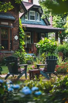 two lawn chairs sitting in front of a house with flowers around them and trees surrounding it