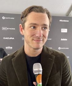 a man holding a microphone in front of a red carpeted wall with an advertisement on it