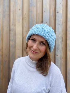 a woman wearing a blue knitted beanie smiles at the camera while standing in front of a wooden fence