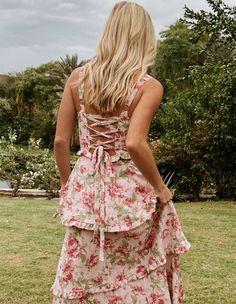 the back of a woman in a floral print dress standing on grass with trees and bushes behind her