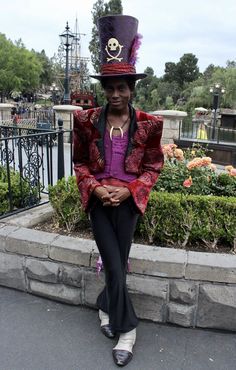 a man wearing a purple top hat and black pants sitting on a stone wall in front of a flower garden