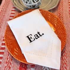 an orange plate with a white napkin that says eat on it sitting on top of a red and white table cloth