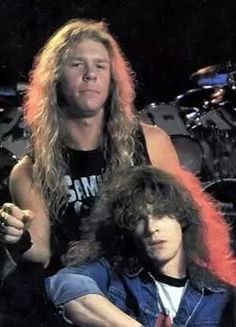 two men sitting next to each other in front of drums and microphones, one with long hair on his head